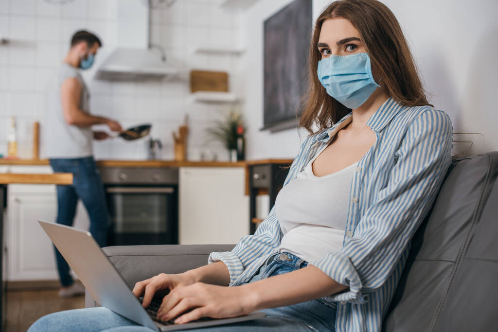 selektiver Fokus der Freiberuflerin in medizinischer Maske, die am Laptop in der Küche arbeitet, in der Nähe ihres Freundes im Hintergrund - Foto, Bild