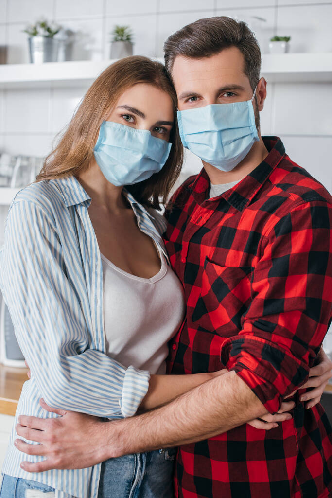 pareja joven en máscaras médicas abrazando y mirando la cámara en la cocina
 - Foto, imagen