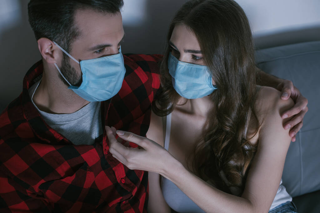young couple in medical masks looking at each other while watching tv  - Photo, Image