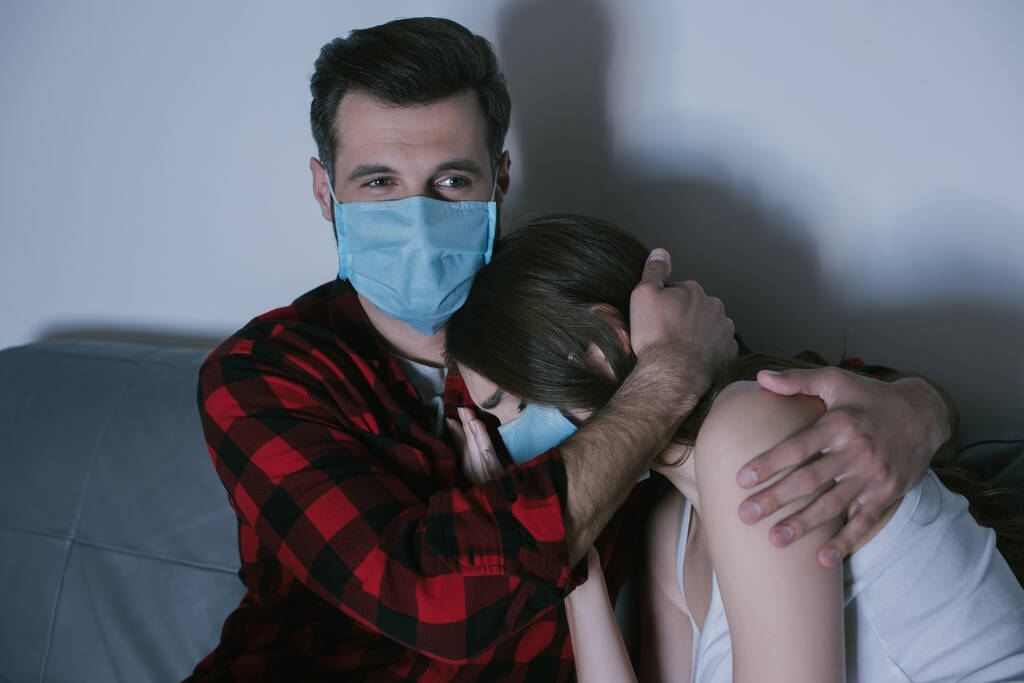 młody para w medyczne maski oglądanie telewizji a człowiek przytulanie zdenerwowany dziewczyna - Zdjęcie, obraz