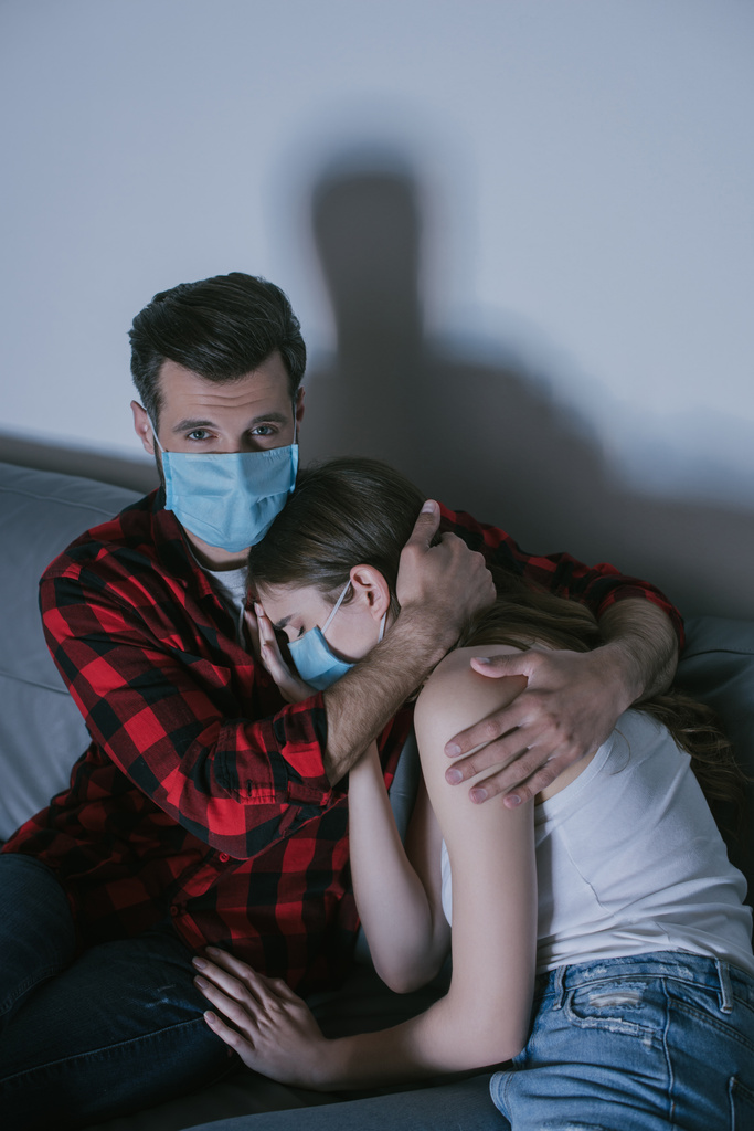 молодая пара в медицинских масках смотрит телевизор, пока мужчина обнимает расстроенную девушку
 - Фото, изображение