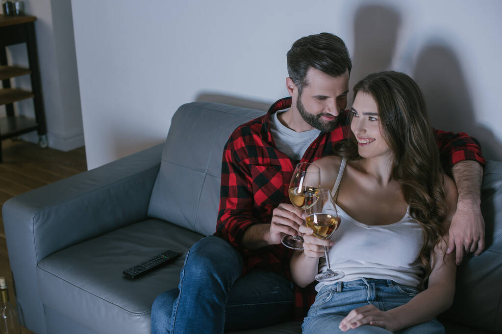 young man embracing happy girlfriend while sitting on sofa with glasses of white wine - Photo, Image