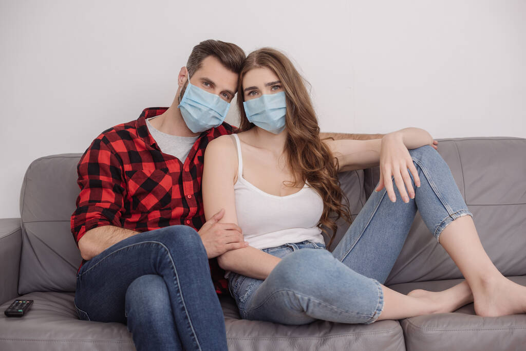 jovem casal deprimido em máscaras médicas sentado no sofá e olhando para a câmera
 - Foto, Imagem