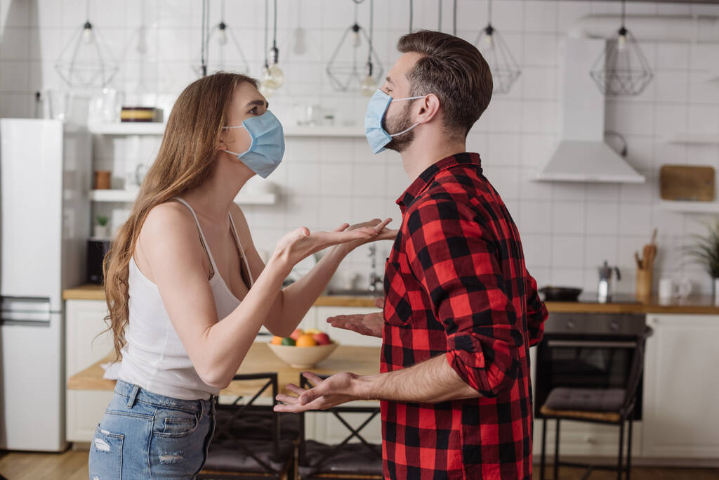 young couple in medical masks quarreling and gesturing in kitchen - Photo, Image