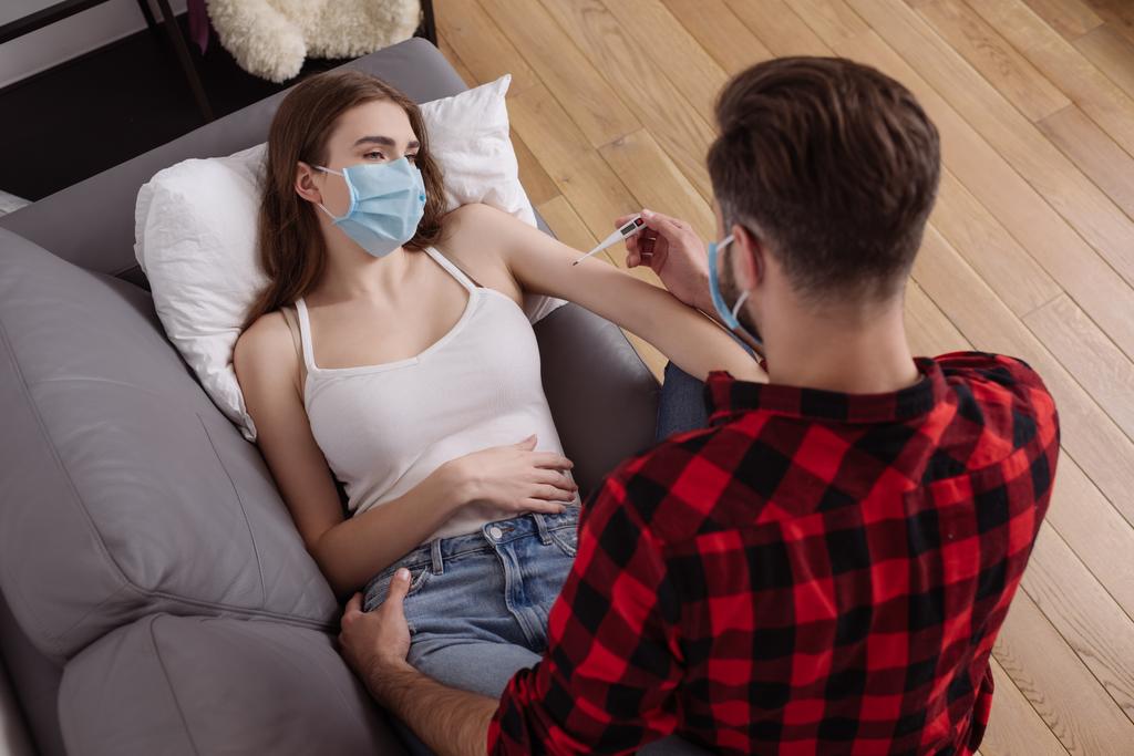 вид сверху на человека, смотрящего на термометр рядом с больной девушкой в медицинской маске лежащей на диване
 - Фото, изображение