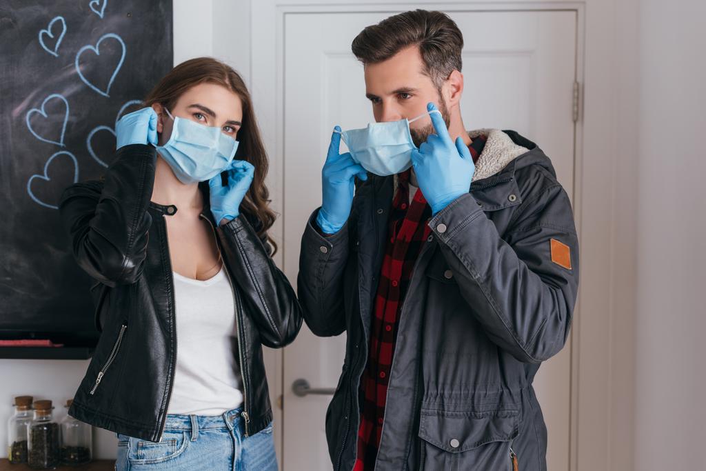 молодая пара в латексных перчатках надевает защитные маски перед уходом из дома
 - Фото, изображение