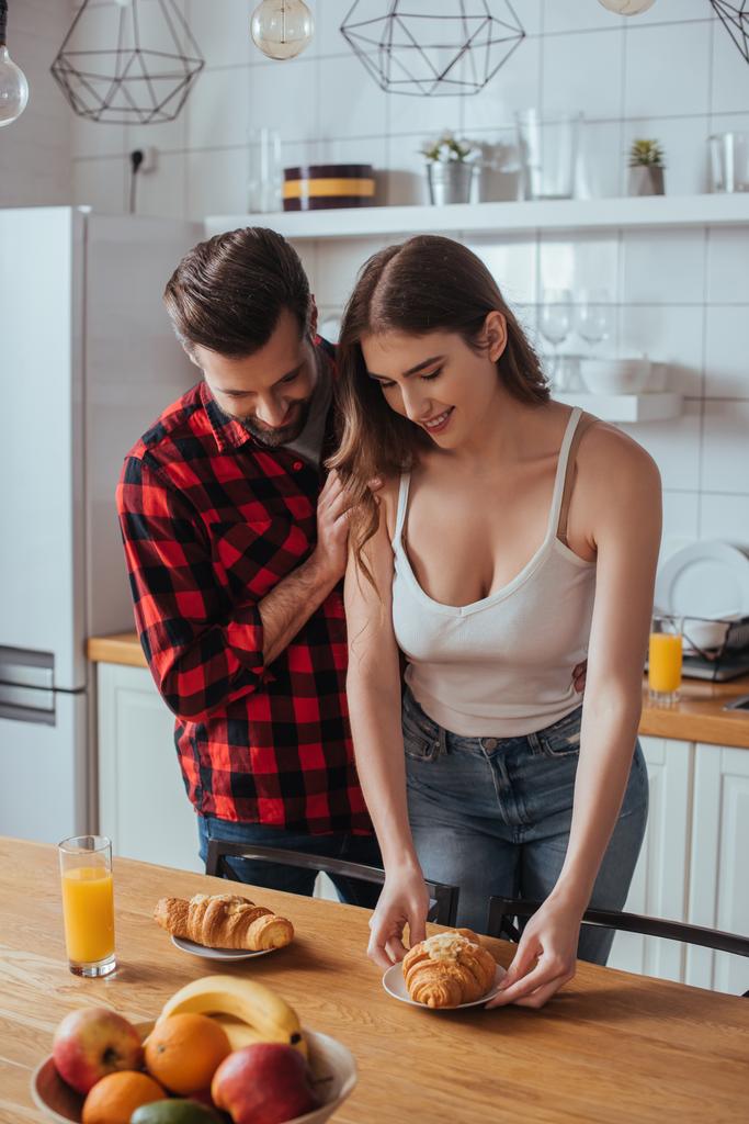 homem bonito tocando ombro de bela namorada segurando prato com croissant saboroso perto de frutas frescas
 - Foto, Imagem