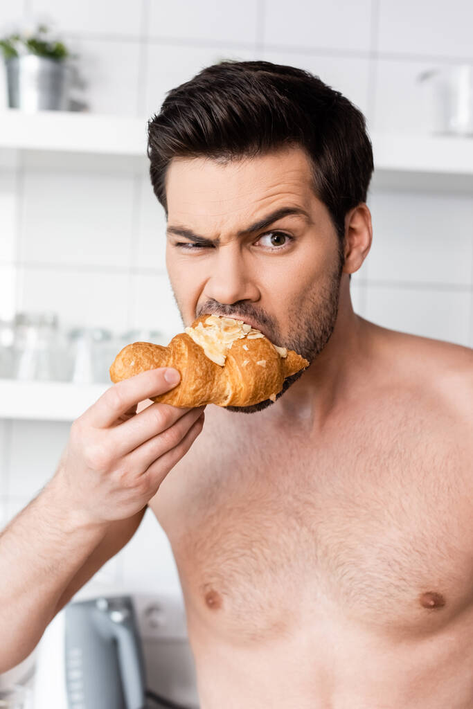 shocked shirtless man eating croissant on kitchen in morning - Photo, Image