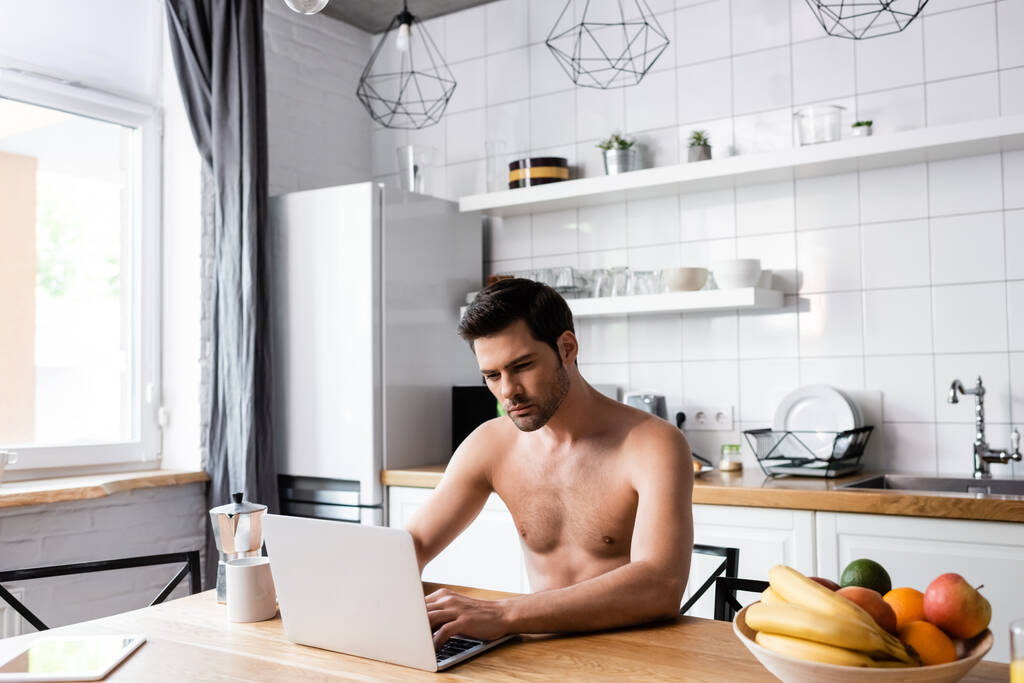 guapo freelancer sin camisa que trabaja en el ordenador portátil en la cocina con frutas y café
  - Foto, imagen