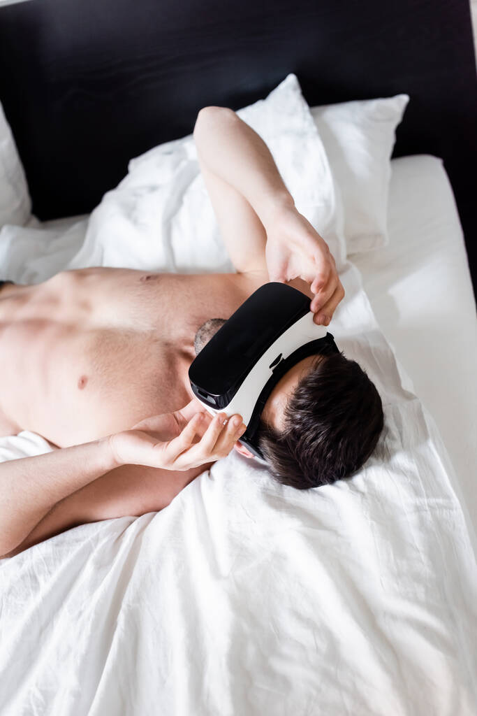 молодой человек без рубашки с помощью гарнитуры виртуальной реальности и лежа на кровати
 - Фото, изображение