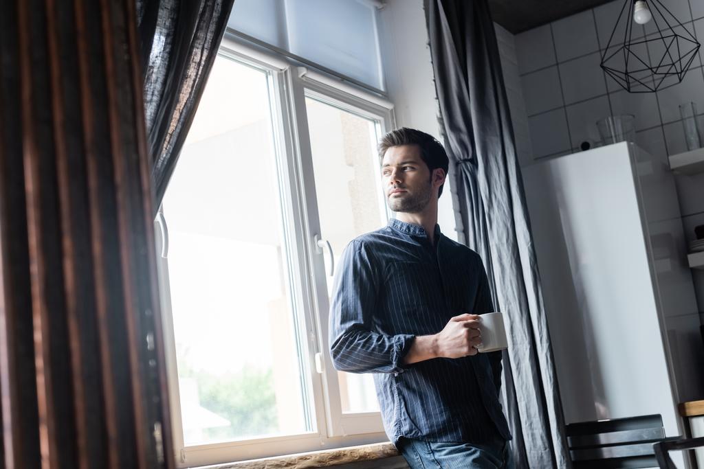 komea mietteliäs mies, joka pitelee kahvikuppia ja seisoo ikkunan lähellä kotona eristäytyneenä - Valokuva, kuva