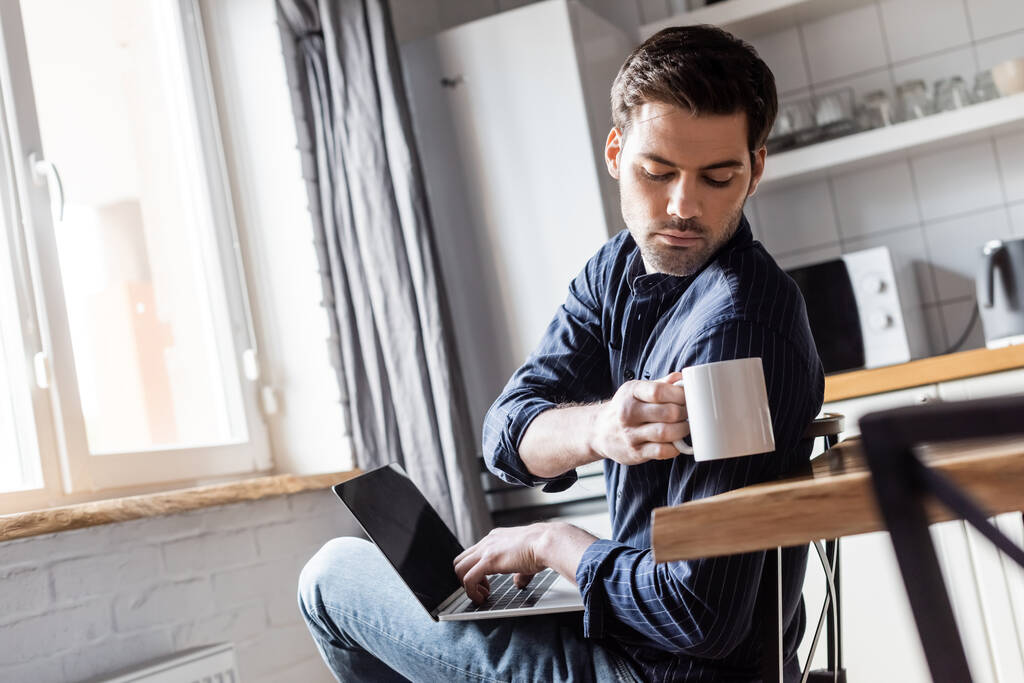 όμορφος άντρας που κρατάει ένα φλιτζάνι καφέ ενώ χρησιμοποιεί λάπτοπ στην κουζίνα κατά τη διάρκεια της καραντίνας   - Φωτογραφία, εικόνα
