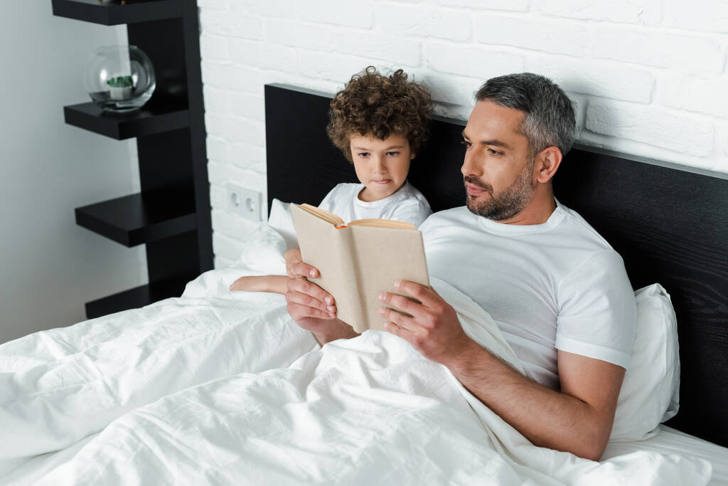 Bärtiger Vater liest Buch neben lockigem Sohn im Schlafzimmer  - Foto, Bild