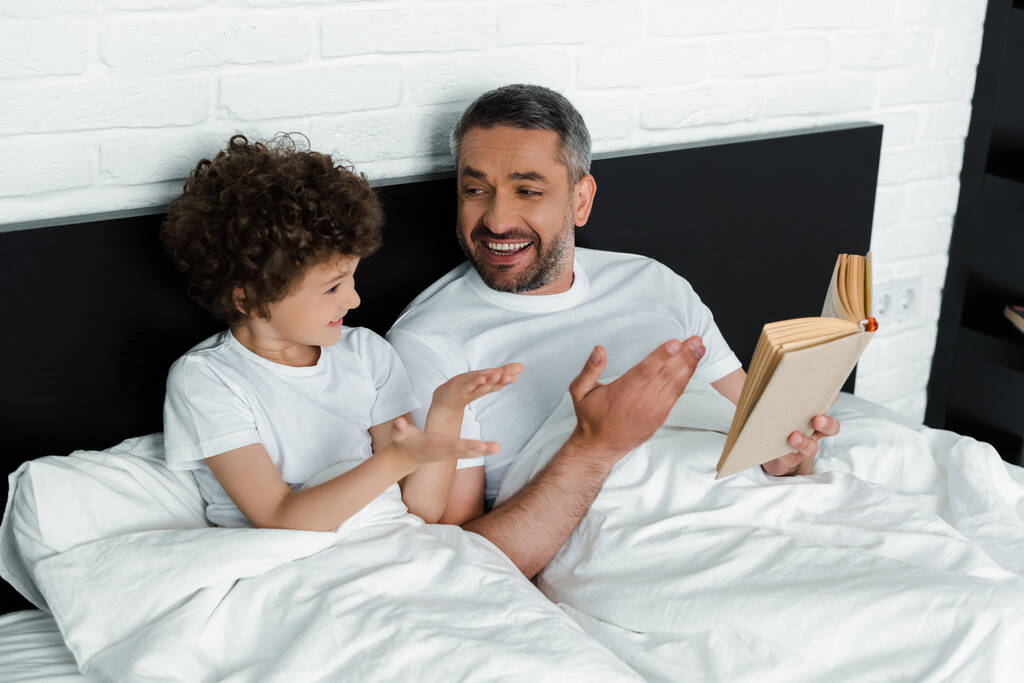 ricci ragazzo gesturing vicino felice padre holding libro in camera da letto
  - Foto, immagini
