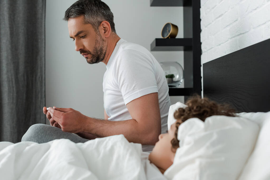 επιλεκτική εστίαση του ανήσυχου πατέρα κρατώντας ψηφιακό θερμόμετρο κοντά άρρωστος γιος στο κρεβάτι  - Φωτογραφία, εικόνα