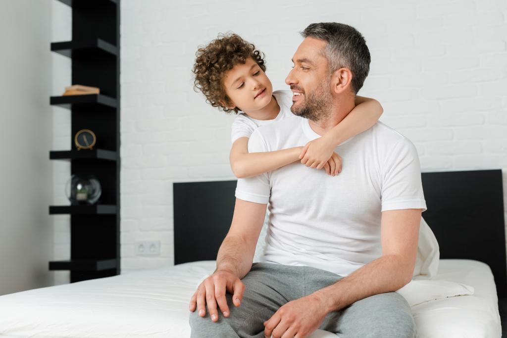 Lockensohn umarmt fröhlichen bärtigen Vater im Schlafzimmer  - Foto, Bild