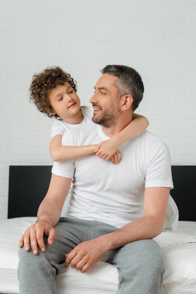Ο σγουρός γιος αγκαλιάζει τον χαρούμενο γενειοφόρο πατέρα και κοιτιέται στην κρεβατοκάμαρα.  - Φωτογραφία, εικόνα