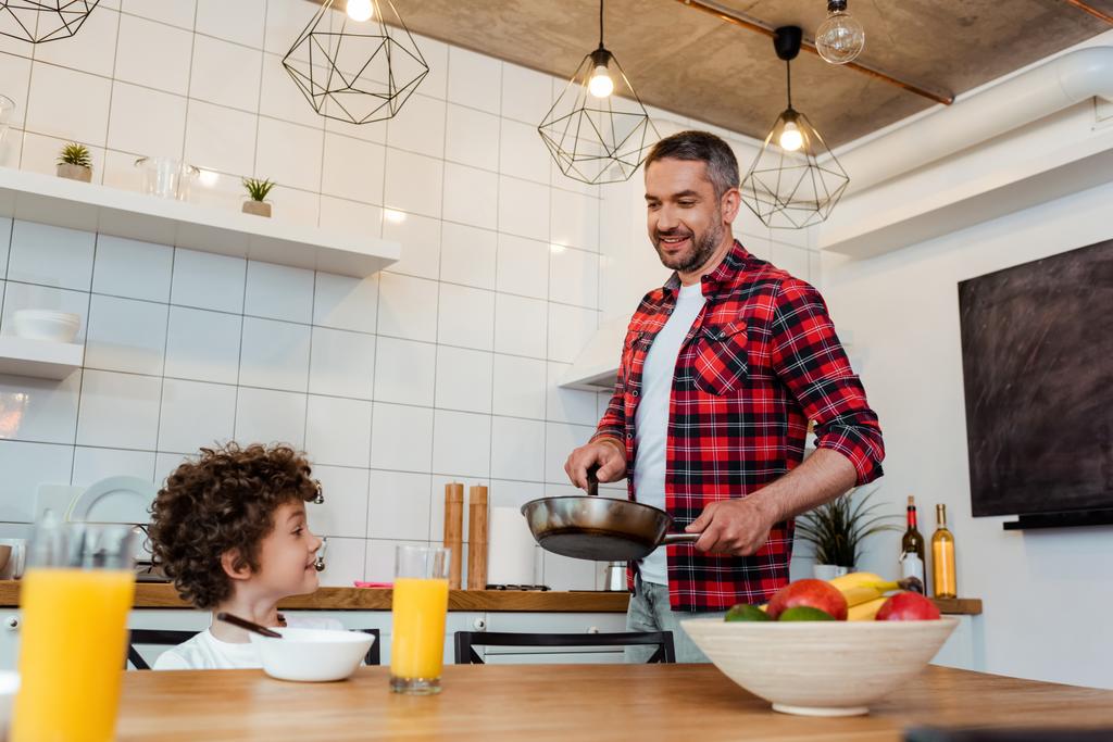 Selektiver Fokus des glücklichen Jungen, der auf den fröhlichen Vater mit der Bratpfanne in der Küche blickt  - Foto, Bild