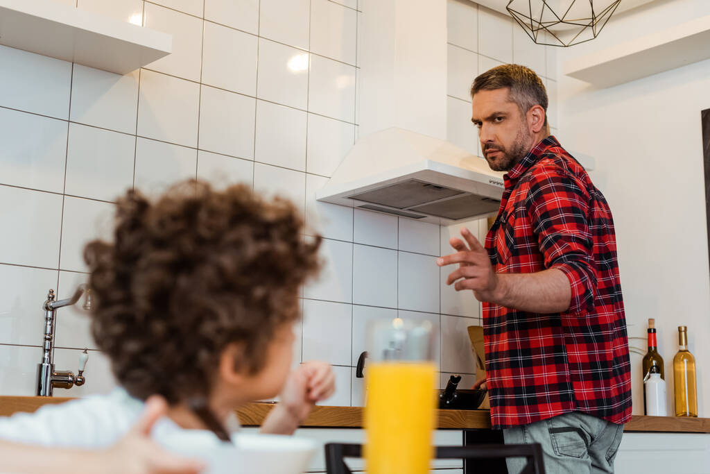 selectieve focus van serieuze vader kijken naar krullende zoon en gebaren in de keuken  - Foto, afbeelding