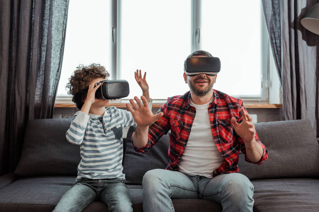 vousatý otec a kudrnatý syn ve sluchátkách virtuální reality gestikulující v obývacím pokoji - Fotografie, Obrázek