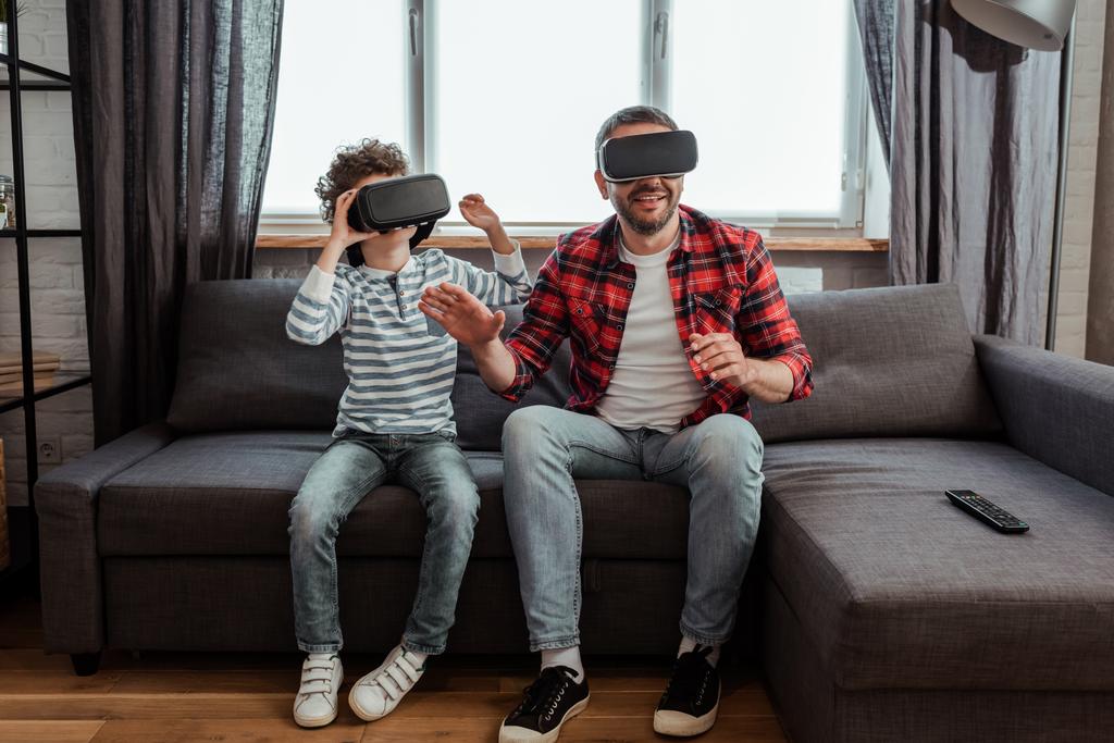 gelukkig vader en krullend zoon in virtual reality headsets zitten in de woonkamer  - Foto, afbeelding