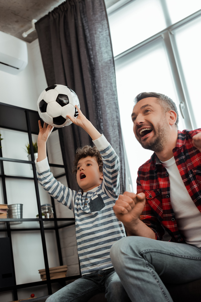 συναισθηματικό παιδί κρατώντας το ποδόσφαιρο, ενώ βλέποντας πρωτάθλημα με ενθουσιασμένο πατέρα - Φωτογραφία, εικόνα