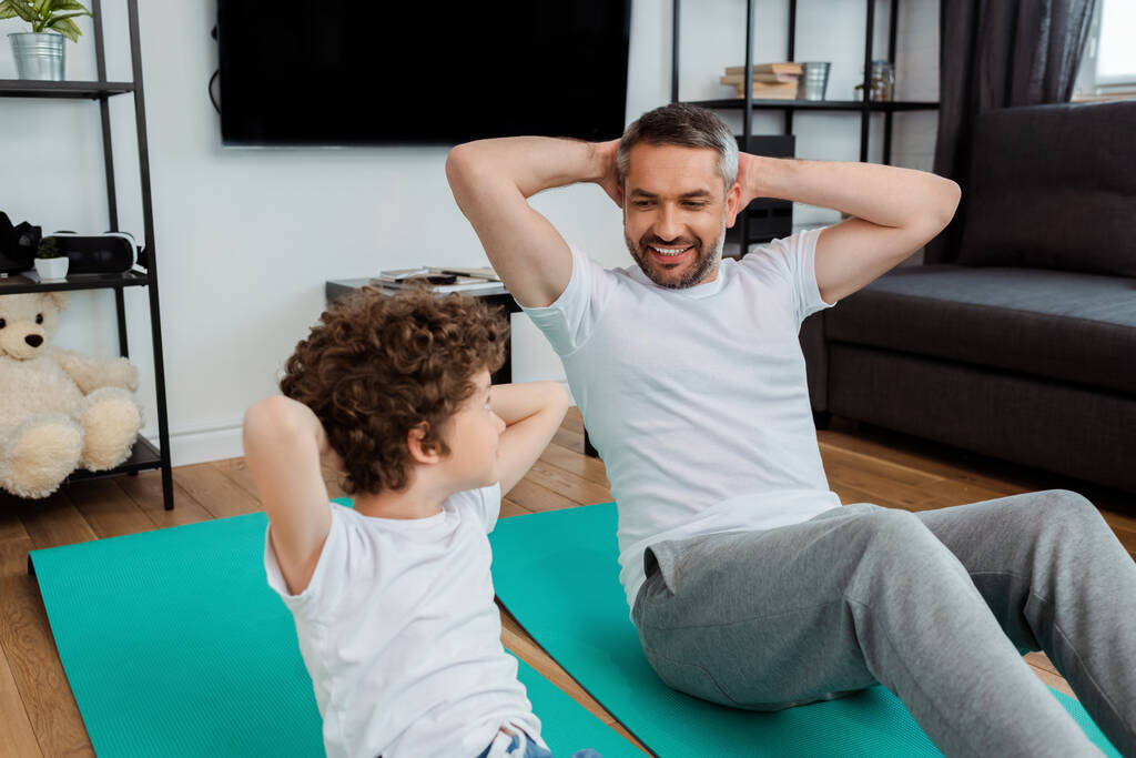 bambino riccio e padre barbuto che si guardano mentre si allenano sui tappetini fitness a casa
 - Foto, immagini