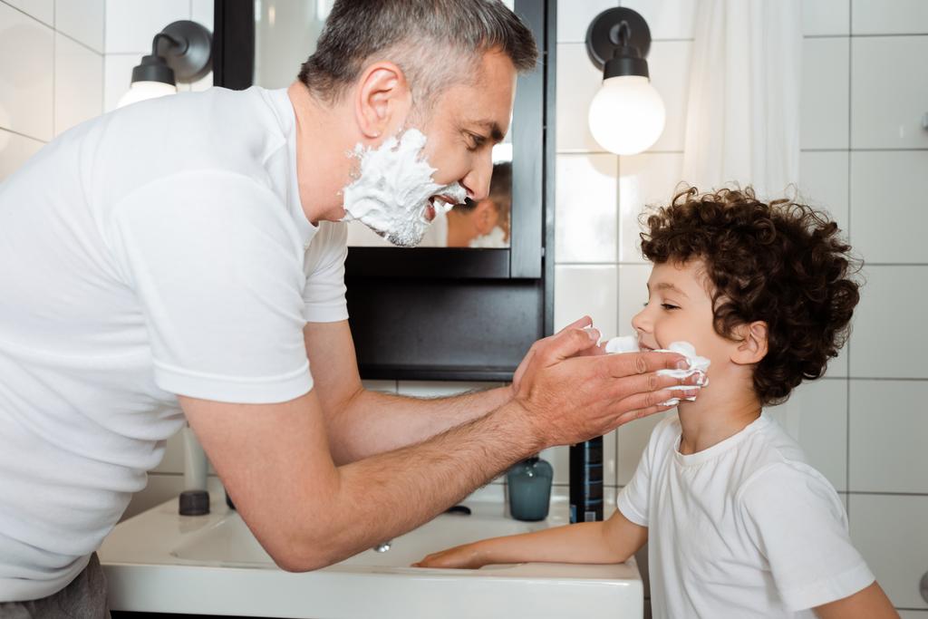 вид збоку щасливого батька, що застосовує піну для гоління на обличчі милого сина у ванній
  - Фото, зображення