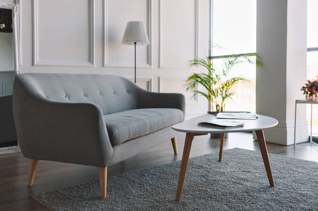 Grauweißes Interieur im skandinavischen Stil. Ein graues Sofa und ein weißer Tisch in einem hellen Raum mit bodenlangen Fenstern. - Foto, Bild
