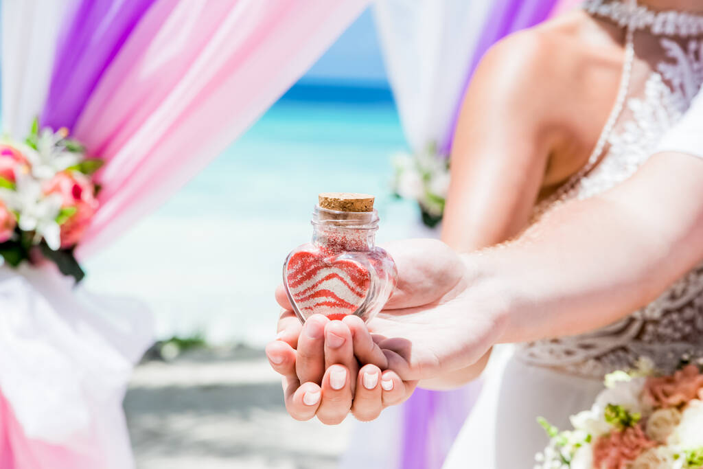 Bruid en bruidegom gieten kleurrijke verschillende gekleurde zand in de kristallen vaas close-up tijdens symbolische nautische decor bestemming bruiloft eenheid ceremonie op zandstrand in de voorkant van de oceaan  - Foto, afbeelding
