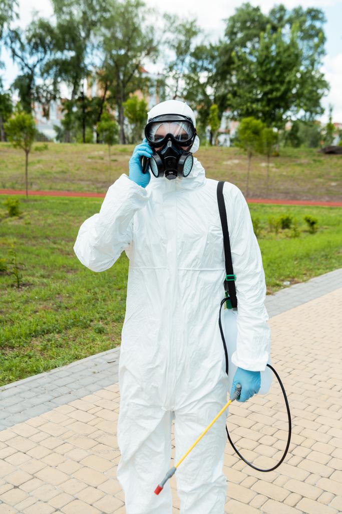 especialista en limpieza en traje de felpudo y respirador sosteniendo bolsa de spray con desinfectante mientras hablaba por celular en el parque durante la pandemia de coronavirus
 - Foto, Imagen