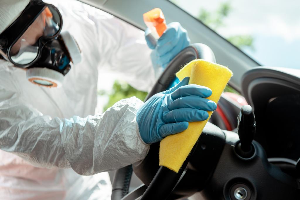 specialist in hazmat suit and respirator cleaning car interior with antiseptic spray and rag during covid-19 pandemic - Photo, Image