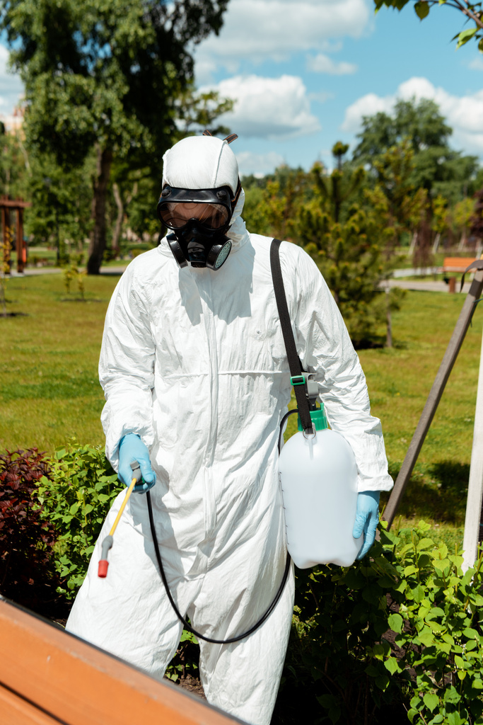 Spezialist in Schutzanzug und Atemschutzmaske desinfiziert Bank im Park während der Coronavirus-Pandemie - Foto, Bild