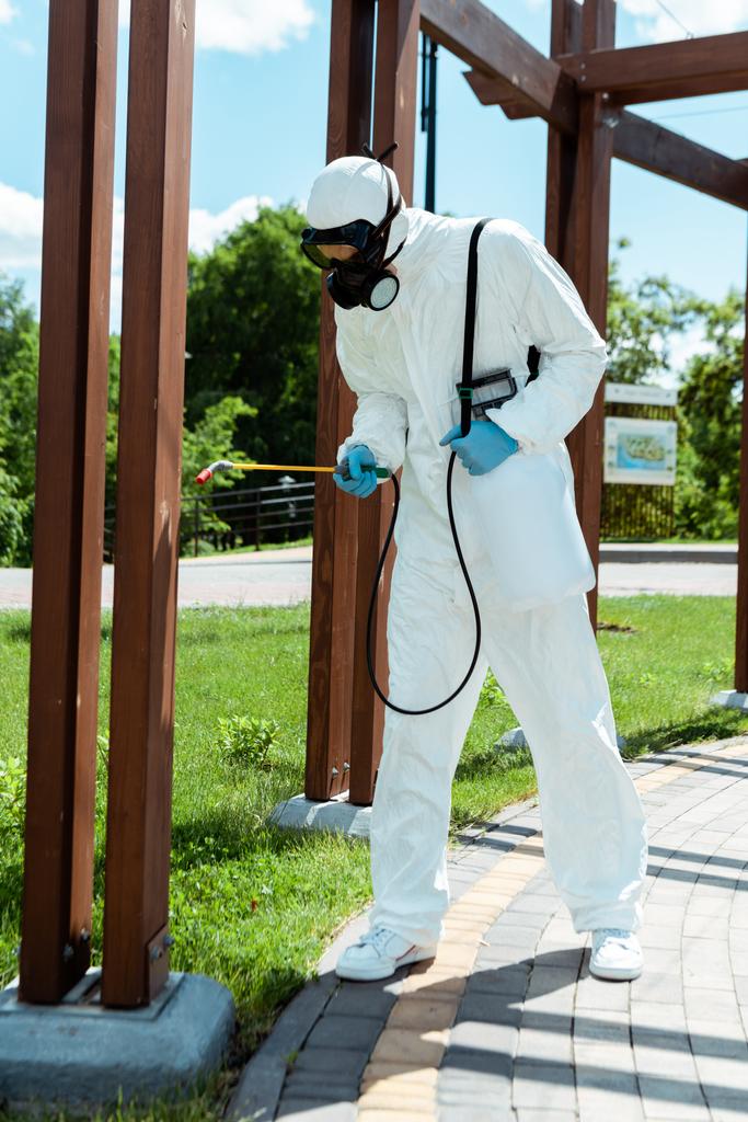 professional specialist in hazmat suit and respirator disinfecting wooden construction in park during coronavirus pandemic - Photo, Image