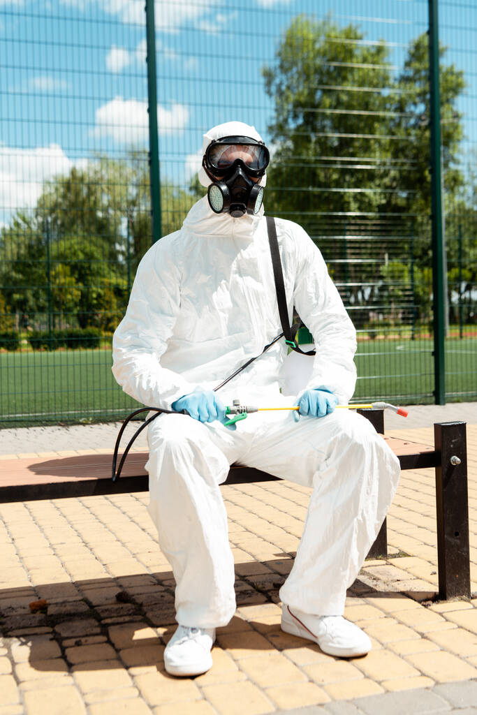 Spezialist in Schutzanzug und Atemschutzmaske sitzt während der Covid-19-Pandemie auf Bank mit Sprühbeutel - Foto, Bild