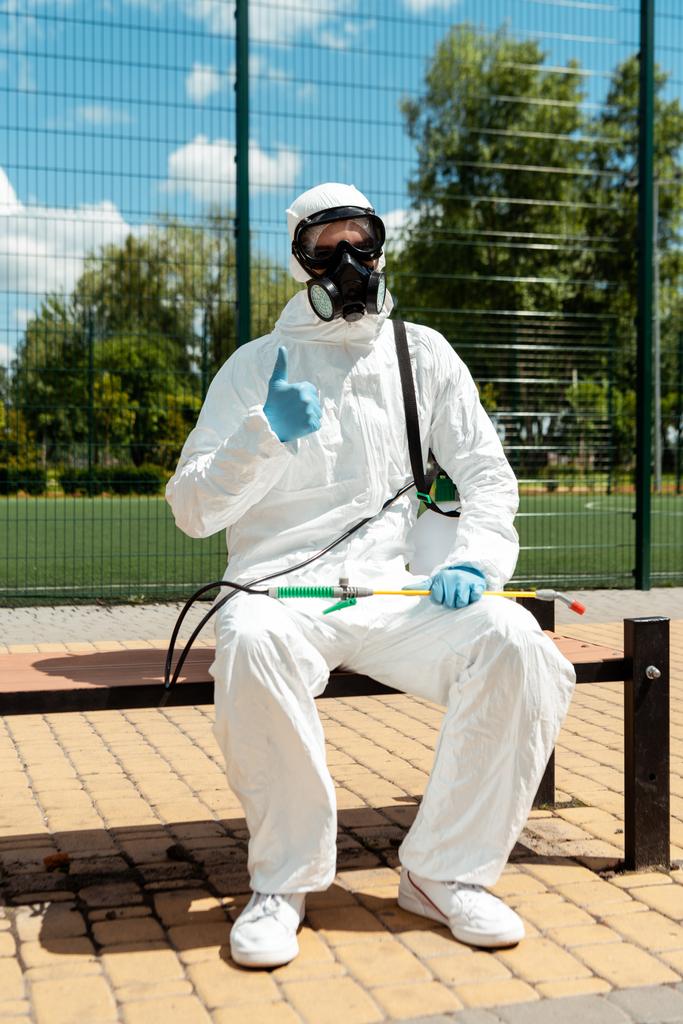 especialista en limpieza en traje de materiales peligrosos y respirador que muestra el pulgar hacia arriba mientras está sentado en el banco con bolsa de aerosol durante la pandemia de covid-19
 - Foto, imagen
