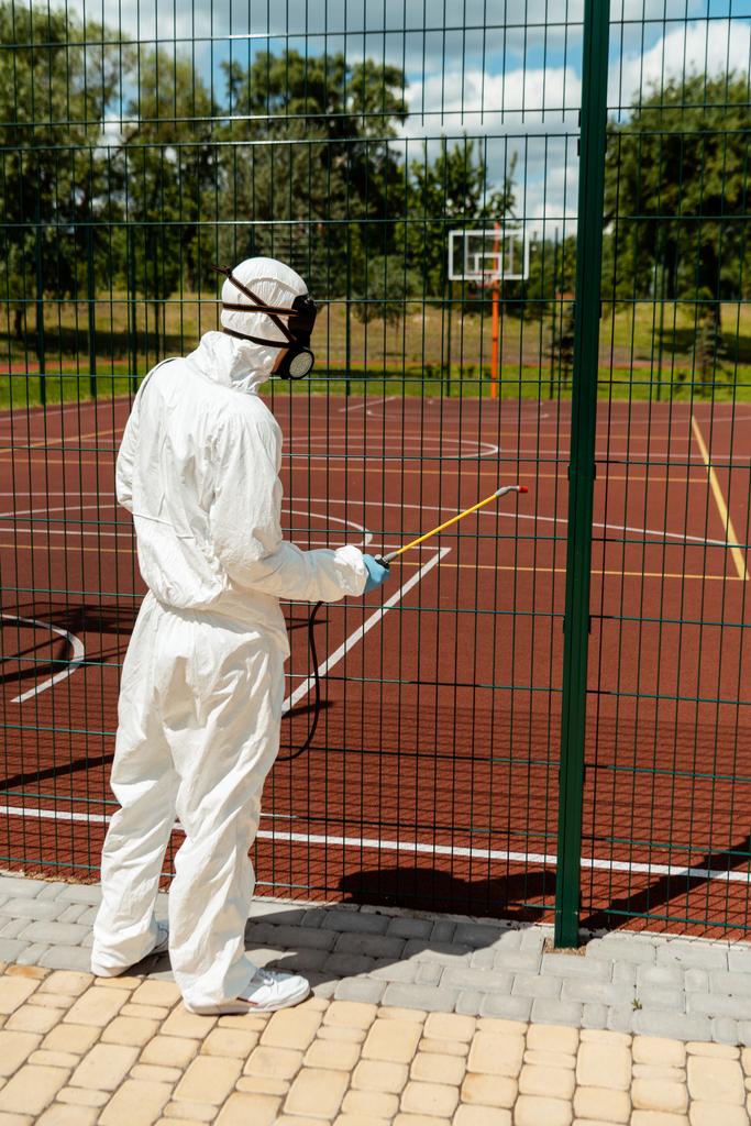 фахівець з маскування костюма та респіратора дезінфікує паркан баскетбольного майданчика в парку під час пандемії ковадла-19
 - Фото, зображення