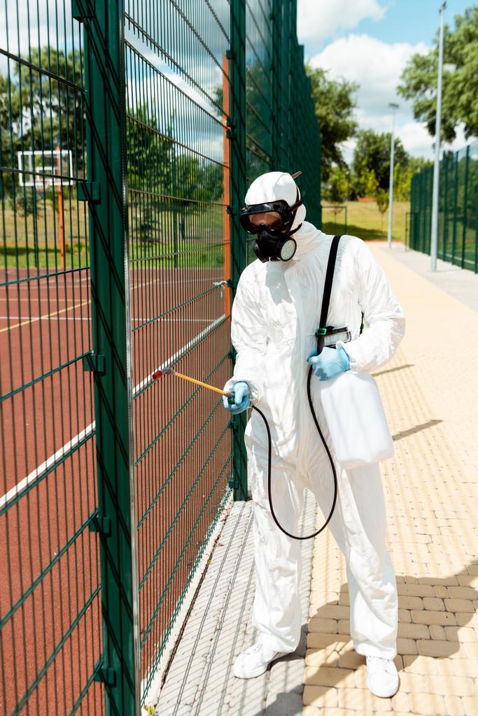 Profi-Spezialist in Schutzanzug und Atemschutzmaske desinfiziert Zaun des Basketballfeldes im Park während der Coronavirus-Pandemie - Foto, Bild