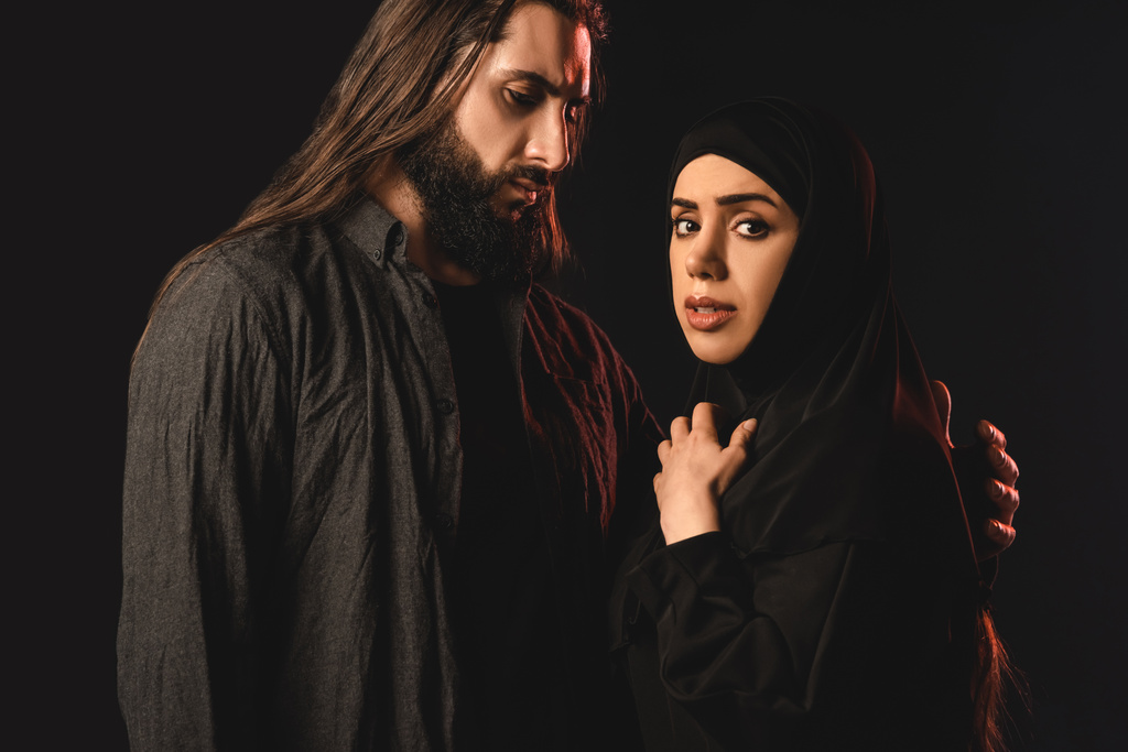 Muslimische Frau im Hidschab blickt in Kamera neben Ehemann, der auf Schwarz isoliert ist  - Foto, Bild