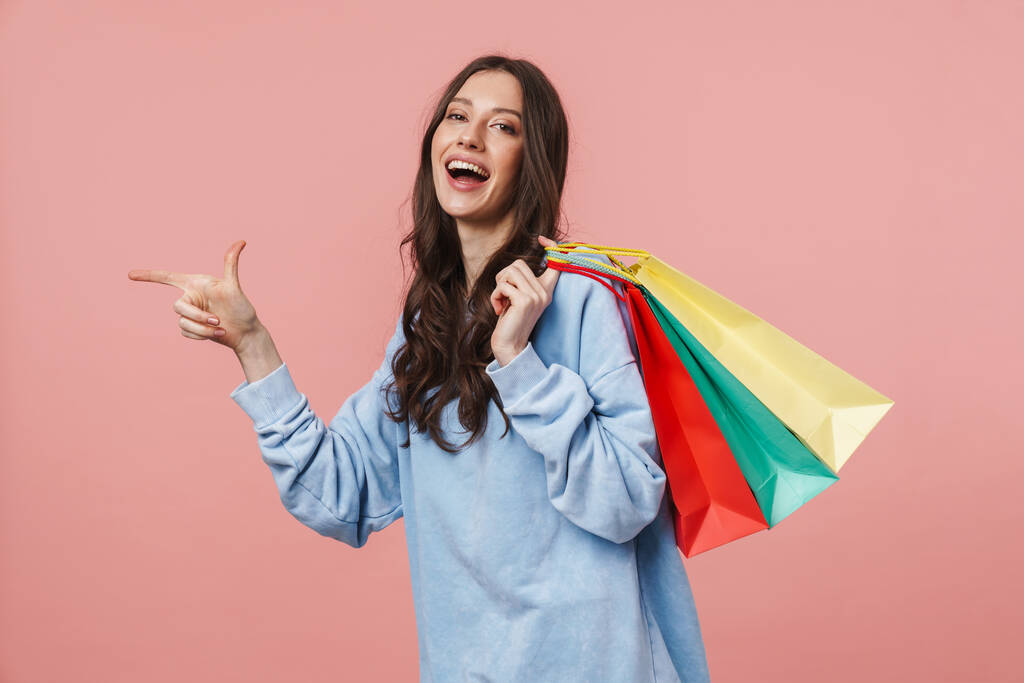 Kép a vonzó fiatal nő hosszú barna haj mutogató ujját, miközben gazdaság bevásárlótáskák elszigetelt rózsaszín háttér - Fotó, kép
