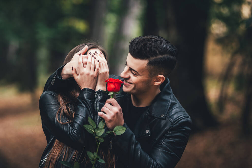 ハンサムなブルネットの男は手で女性の目をカバーし、彼女の赤いバラを与え、秋の公園で. - 写真・画像