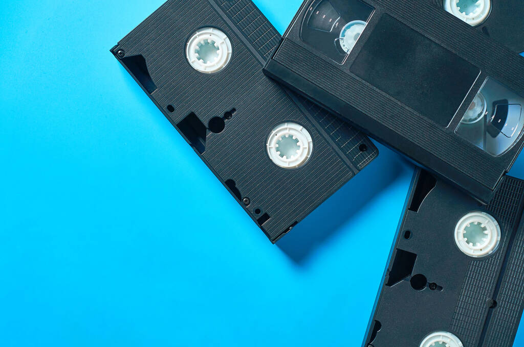 Rozrzucone wiele czarnych starych plastikowych kaset wideo vhs leży na niebieskim biurku. Koncepcja lat 90. Odbiór. Układy płaskie - Zdjęcie, obraz