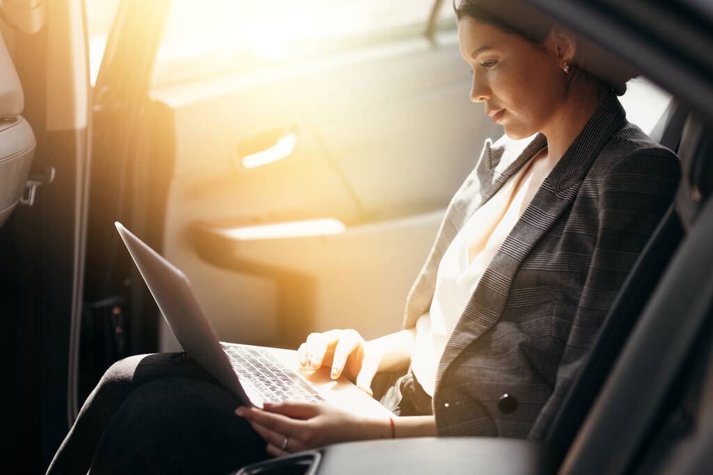 Businesswoman in a jacket sits in a car in the back seat and works at a laptop checking work mail - Photo, Image