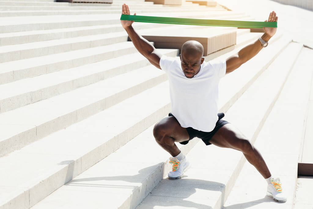 Dark-skinned African professional athlete squats with elastic athletic tape. Training outdoor for strengthening the biceps and quads. - Photo, Image