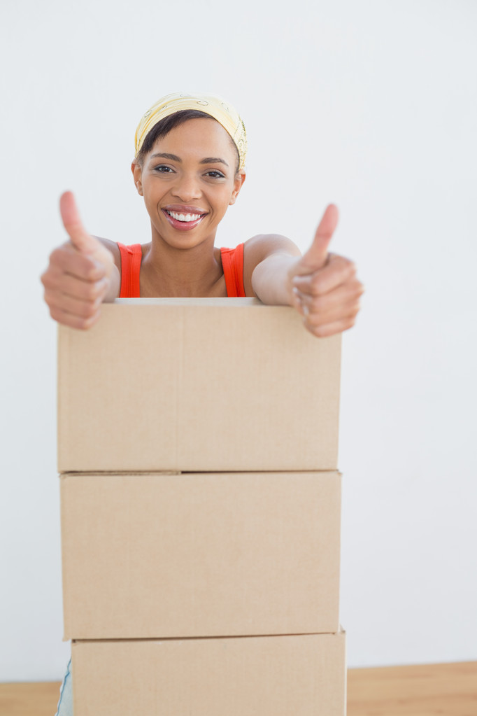 Woman gesturing thumbs up with stack of boxes in a new house - Photo, Image