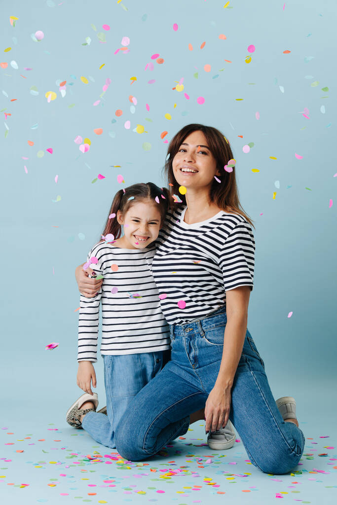 Two fashion lovers, mom and daughter wearing same clothes. They have jeans and black and white striped shirt on them. Standing on their knees, smiling, celebrating under a confetti shower. - Photo, Image