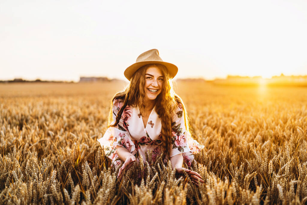 長い巻き毛とそばかすの顔を持つ若い女の子,帽子で,花のプリントと薄い白いドレスで,小麦畑に立って,カメラのポーズ,背景の日没で - 写真・画像