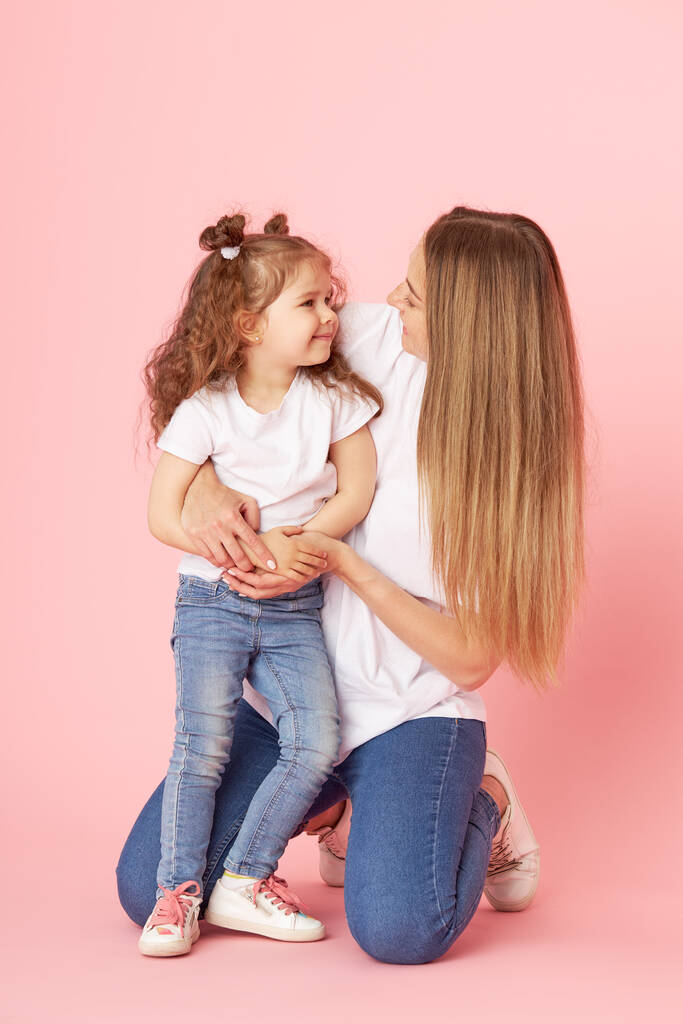 Äiti ja tytär valkoisissa t-paidoissa ja farkuissa leikkivät ja halaavat vaaleanpunaisella taustalla. Läheisistä huolehtiminen. Onnellinen äitiys
 - Valokuva, kuva