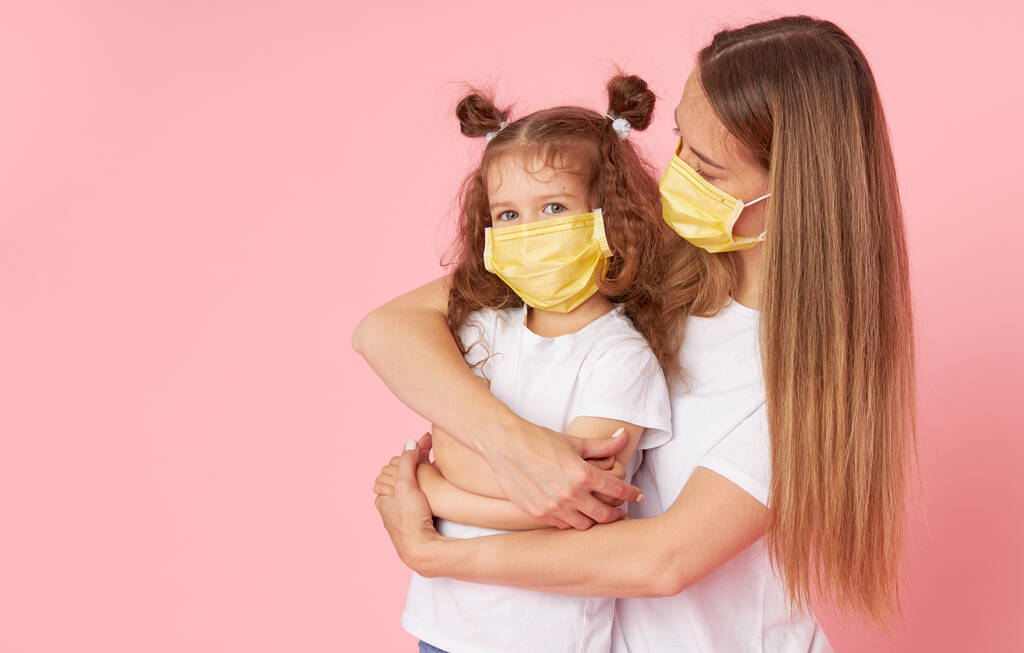 Zorgen voor geliefden. Jonge moeder knuffelt kleine dochter in beschermende medische maskers tijdens Covid-19 pandemieën. Studio roze achtergrond - Foto, afbeelding