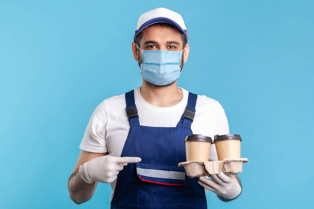 Delivery service. Optimistic courier man in overalls and mask pointing at coffee, wearing safety gloves offering drinks in disposable cups and looking at camera with smile. indoor studio shot isolated - Photo, Image